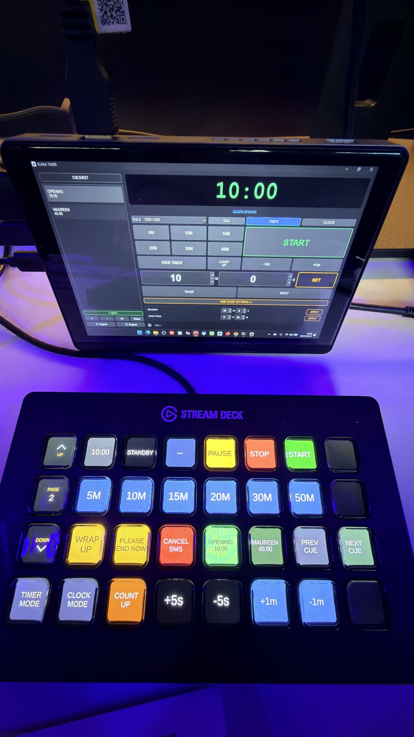 Live show set-up — KUMA Timer running on a tablet in the background showing the cuesheet and 10:00 countdown, with an Elgato Stream Deck in the foreground laid out with KUMA buttons (START / PAUSE / STOP, 5M-50M presets, WRAP UP / PLEASE END NOW / CANCEL SMS, ±1m / ±5s and timer-mode controls).